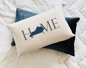 May include: A white pillow with the word "HOME" printed in gray with the state of Kentucky in blue in the middle of the word. The pillow is on top of a dark blue pillow.