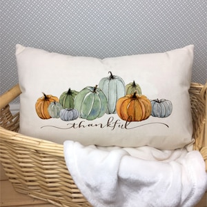 May include: Cream-colored rectangular pillow featuring a watercolor design of pumpkins in shades of orange, green, and blue. The word "thankful" is written in a script font below the pumpkins. The pillow is placed in a woven basket.
