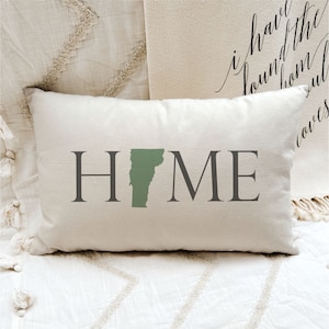 May include: A white pillow with the word "HOME" in black text and a green outline of the state of Vermont. The pillow is on a white bedspread.