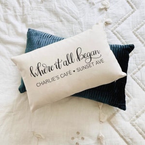 Puede incluir: Dos cojines decorativos sobre una superficie acolchada blanca. Uno es blanquecino con el texto "& Where it all began" y "Charlie's Caf&eacute; Sunset Ave". Los otros cojines son de color verde azulado oscuro.