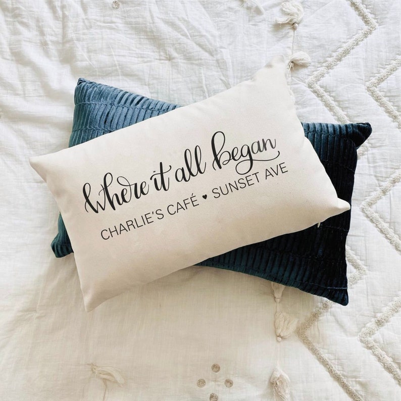 Puede incluir: Dos cojines decorativos sobre una superficie acolchada blanca. Uno es blanquecino con el texto "& Where it all began" y "Charlie's Caf&eacute; Sunset Ave". Los otros cojines son de color verde azulado oscuro.