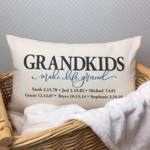 以下が含まれることがあります： 黒と青の文字で「GRANDKIDS make life grand」と書かれた白い枕。テキストの下には、孫の名前と生年月日が記載されています。