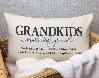 Puede incluir: Almohada blanca con el texto "GRANDKIDS make life grand" en letras negras y azules. Debajo del texto aparecen los nombres de los nietos y sus fechas de nacimiento.