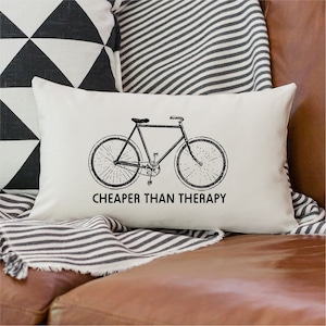 May include: White throw pillow with a black and white illustration of a bicycle and the text "Cheaper than therapy".