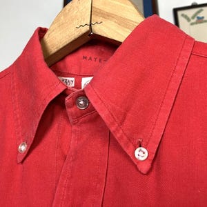 May include: Close-up of a red button-down shirt with a classic collar. The shirt is hanging on a wooden hanger. The collar has a button on each point. The shirt is made of a solid red fabric.