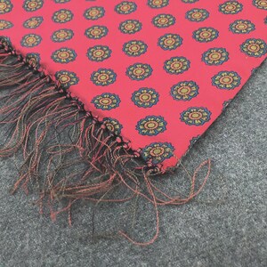 May include: A rectangular fabric scarf in a vibrant coral pink color featuring a repeating pattern of small circular floral motifs in dark blue and gold. The edge of the scarf is finished with long, thin, loose fringe threads in mixed black and red tones.