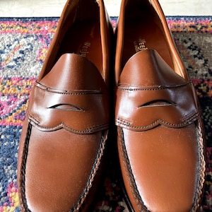 May include: A pair of brown leather loafers with a penny slot detail. The loafers have a black leather trim and a black leather sole.