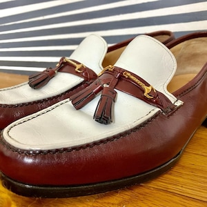 May include: A pair of two-tone leather loafers. The shoes feature a white vamp and a burgundy body with a gold-colored accent and tassel detail. The shoes are on a wooden surface.