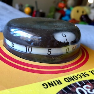 以下が含まれることがあります： 白の文字盤と赤いアクセントが付いた黒いキッチンタイマー。タイマーには、0、5、10、15の数字が書かれたダイヤルがあります。タイマーには「0 half」という文字が見えます。タイマーは、黄色と赤の背景に置かれており、「SIX SECOND RING 30」という文字が見えます。