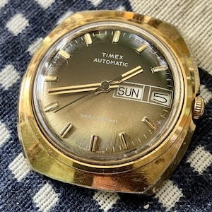 Op de afbeelding: Een vintage goudkleurig Timex Automatic horloge met een bruine wijzerplaat. Het horloge heeft gouden wijzers en markeringen, een dag-datumweergave en de tekst "TIMEX AUTOMATIC" en "WATER RESISTANT". Het horloge ligt op een blauw-wit geruit oppervlak.