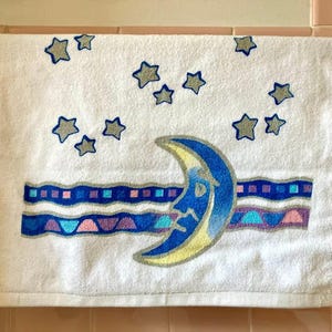 May include: White bath towel with a blue crescent moon and stars printed on it. The moon has a yellow crescent shape and a face. The stars are silver and scattered across the towel. There are blue, purple, and pink stripes below the moon.
