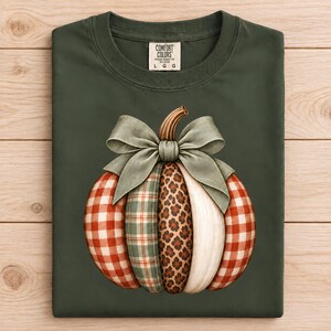 May include: A dark olive green t-shirt featuring a large graphic of a pumpkin. The pumpkin is composed of vertical segments with red and white gingham, green plaid, leopard print, and solid cream patterns, topped with a sage green bow.