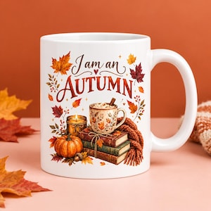 May include: A white ceramic mug featuring a colorful autumn-themed illustration. The design includes the text 'I am an AUTUMN' surrounded by orange and red maple leaves, a pumpkin, a lit candle, a stack of books, and a mug of hot cocoa.
