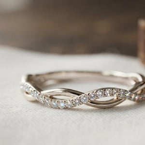 Può includere: Un anello color argento lucido caratterizzato da un design a fascia intrecciata. Un filo della fascia è incastonato con una fila continua di piccole pietre trasparenti a taglio rotondo che catturano la luce, mentre l'altro filo rimane liscio.