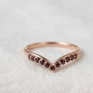 Pode incluir: Um anel em forma de V feito de metal em tons rosados, apresentando uma série de pequenas pedras preciosas granadas redondas e vermelho-escuras cravadas ao longo da curva frontal da banda. O design segue uma silhueta em chevron polida.