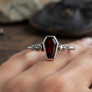 Puede incluir: Un anillo de plata con una piedra preciosa de color rojo oscuro en forma de ataúd en el centro. La banda presenta pequeños motivos de calaveras esculpidas a ambos lados del engaste, unidos por intrincados adornos metálicos.