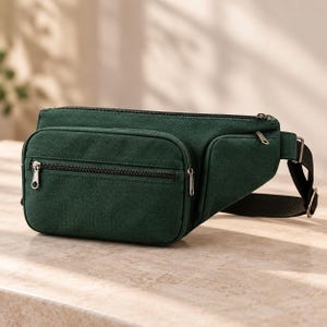 May include: A dark forest green canvas bum bag featuring a large main zipped compartment, a smaller front zipped pocket, and a black adjustable strap with a metal buckle. The bag has a structured rectangular shape with rounded corners.