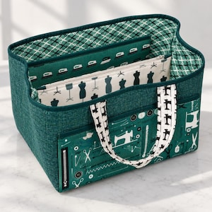 May include: A teal fabric storage tote featuring a sewing-themed print. The exterior displays sewing machines, scissors, and zippers. The interior lining shows a pattern of irons and scissors, while a central divider features dress forms.