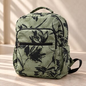 Könnte beinhalten: Ein salbeigrüner Rucksack mit einem schwarzen, pinselstrichartigen Blumenmuster. Der Rucksack verfügt über einen Tragegriff, ein großes Hauptfach mit Reißverschluss, eine kleinere Fronttasche mit Reißverschluss sowie Seitentaschen mit schwarzem Rand.