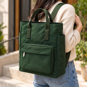 May include: A forest green rectangular backpack featuring two top carry handles, adjustable shoulder straps, and a large front zippered pocket. The bag includes side pockets and is constructed from a solid-colored fabric with matching webbing.