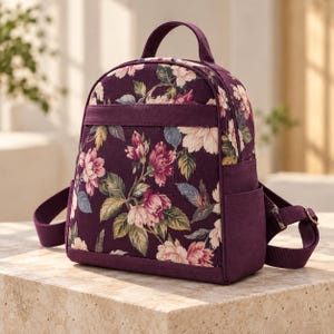 May include: A small backpack featuring a deep purple fabric base with a prominent floral pattern in shades of pink, cream, and green. The bag includes a top handle, adjustable shoulder straps, a horizontal front band, and side pockets.