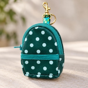 May include: A miniature backpack-shaped pouch in dark green with a light blue polka dot pattern. It features a teal zipper closure, a horizontal front pocket detail, and a gold-toned metal lobster clasp attached to a small floral fabric loop.