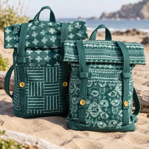 Pode incluir: Duas mochilas de fecho enrolável em tons de verde escuro. A mochila da esquerda apresenta um padrão de cruzes brancas e um design de grelha. A da direita exibe uma faixa geométrica e uma estampa floral. Ambas têm alças e botões amarelos.