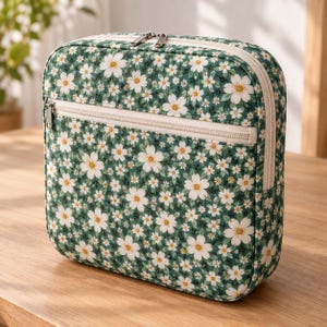 Könnte beinhalten: Eine quadratische Aufbewahrungstasche mit dunkelgrünem Hintergrund und einem wiederkehrenden Muster aus weißen Blumen und kleinen grünen Blättern. Die Tasche verfügt über einen beigen Reißverschluss oben und eine Fronttasche mit passendem Reißverschluss.