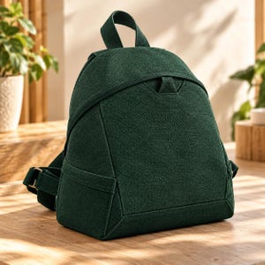 May include: A dark forest green backpack with a textured fabric finish. The bag features a rounded top handle, adjustable shoulder straps with metal buckles, and a structured, tapered design with a horizontal seam detail across the front panel.