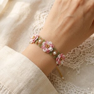 May include: A delicate bracelet featuring three crocheted flowers in shades of pink and white with yellow centers, accented by small green crocheted leaves and round white pearls. The floral design is connected by a thin gold-toned metal chain.