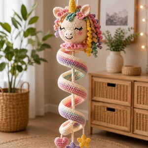 Op de afbeelding: Een gehaakte eenhoorn hangdecoratie met een crèmekleurig hoofd, een gele hoorn, roze wangen en een veelkleurige krullende manen. Een lange, spiraalvormige staart in pastelkleurige regenboogstrepen hangt eronder, eindigend in kleine hart- en sterbedels.