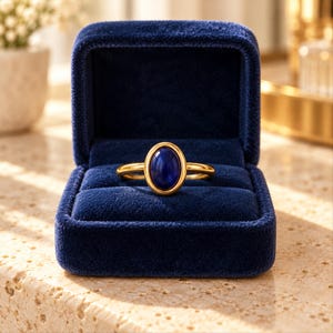 May include: A gold ring featuring a smooth, oval-shaped deep blue stone set in a polished gold bezel. The ring is displayed centered within a dark blue velvet jewelry box with a hinged lid, showcasing the contrast between the metal and stone.
