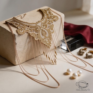 May include: A gold lace collar necklace featuring intricate embroidery and a series of dangling gold chains with pointed metal spikes. The piece includes long, layered gold chains extending from the sides and is accented by three white pearls.