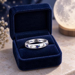 May include: A silver-toned band ring featuring inlaid celestial motifs including crescent moons and star shapes, accented with small round blue and clear stones, presented inside a deep blue velvet-lined jewelry box.