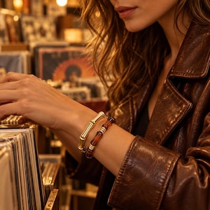 May include: Two bracelets worn on the wrist. One bracelet is made of polished gold-tone metal with a segmented bamboo-like design. The second bracelet features dark brown, translucent, cylindrical beads separated by small gold-tone metal rings.
