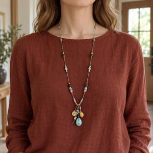 Puede incluir: Un collar largo con un cordón gris ensartado con varias cuentas en tonos turquesa, ámbar y gris oscuro. La sección del colgante consiste en un grupo de dijes, que incluye una pequeña moneda dorada, una cuenta amarilla y una lágrima turquesa.