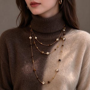 May include: A multi-strand gold-tone necklace featuring a delicate chain interspersed with smooth cream-colored beads and dark brown, polished stone beads. The long, layered design rests against a two-toned brown and beige turtleneck sweater.