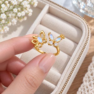 May include: A gold-toned open ring featuring two butterfly motifs. One butterfly is accented with an iridescent oval stone, while the other displays an iridescent oval stone paired with two small, clear, round sparkling stones.