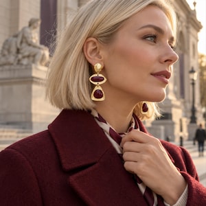 May include: A pair of gold-toned drop earrings featuring a textured circular stud, a horizontal oval-shaped deep red stone, and a triangular-shaped deep red stone. The earrings have a polished metal finish and are worn on the earlobes.