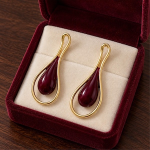 May include: A pair of gold-toned earrings featuring a smooth, teardrop-shaped burgundy enamel center encased within a continuous, open-loop gold metal frame. The earrings are presented inside a deep red velvet-lined jewelry box with cream padding.