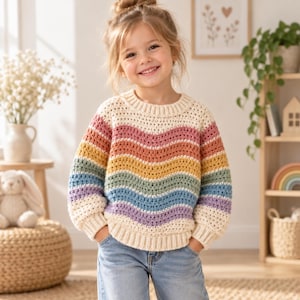 May include: A long-sleeve crochet sweater featuring a cream-colored base with horizontal wavy stripes in shades of pink, orange, yellow, green, blue, and purple. The sweater has a ribbed crew neckline and ribbed cuffs, paired with light blue denim jeans.
