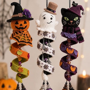 May include: Three hanging crochet ornaments featuring a jack-o'-lantern in a witch hat, a white ghost in a top hat, and a black cat in a purple hat. Each figure has a long, spiraling tail decorated with small crocheted motifs and dangling beads.