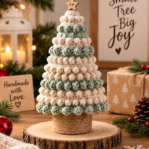 May include: A crocheted Christmas tree decoration featuring alternating horizontal bands of cream and sage green yarn with a textured bobble stitch pattern. The tree is topped with a small wooden star and sits on a cylindrical base wrapped in twine.