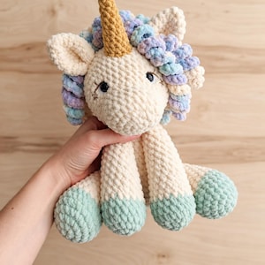 Puede incluir: Un peluche de unicornio tejido a ganchillo en color crema con un cuerno dorado texturizado, una melena de lana rizada en tonos azul pastel, morado y crema, y pezuñas verde menta. El unicornio tiene ojos bordados oscuros con pestañas y estrellas.