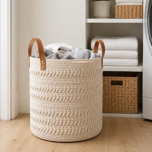May include: A cylindrical laundry basket made of cream-coloured woven rope with a textured herringbone pattern. The basket features two tan faux leather handles attached with gold-toned metal rivets and is filled with various folded clothing items.