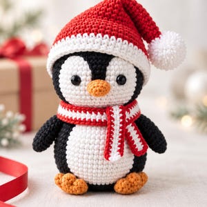 Op de afbeelding: Een gehaakt pinguïnfiguurtje met een zwart-wit lichaam, oranje snavel en voeten, met een rood-witte kerstmuts met een witte pompon en een bijpassende rood-wit gestreepte sjaal. Het speelgoed heeft zwarte kraaloogjes en een textuur van garen.