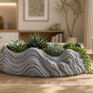 May include: A grey, oval-shaped planter with a textured, wavy surface design. The planter contains various green succulents and a trailing plant. A measurement line indicates a width of 21.3 cm. The planter sits on a wooden surface.