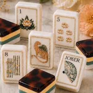 Puede incluir: Una colección de fichas de mahjong rectangulares con caras de color crema que muestran ilustraciones de un loto, un leopardo, bambú y un abanico, junto con fichas con los números 1, 5 y 8, y una ficha de Joker. Algunas fichas tienen una parte superior de carey.