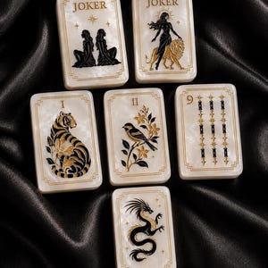 Può includere: Sei carte da gioco rettangolari con finitura bianco perlato e bordi dorati. Due carte sono etichettate JOKER, una con due figure, una con una figura e un leone. Altre carte raffigurano una tigre, un uccello su un ramo, il numero 9 e un drago.