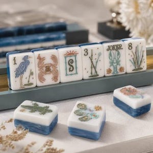 May include: A set of rectangular mahjong tiles with white faces and blue marbled backs. The tiles feature engraved illustrations including a heron, crab, sun, bamboo, lobster labeled JOKER, number 8, alligator, flowers, and the letter S.